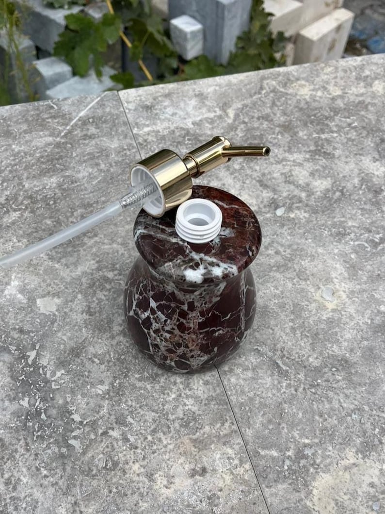 Puede incluir: Un dispensador de jab&oacute;n decorativo con un patr&oacute;n similar al m&aacute;rmol en tonos burdeos y blanco. El dispensador tiene una bomba dorada y un tubo de pl&aacute;stico transparente. La bomba est&aacute; desenroscada, revelando una abertura de pl&aacute;stico blanco.