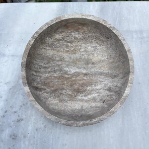 May include: A round, stone bowl with a textured, marbled appearance. The bowl is a light gray color with darker gray and brown veining throughout. The bowl's interior is smooth and the exterior has a rougher texture.