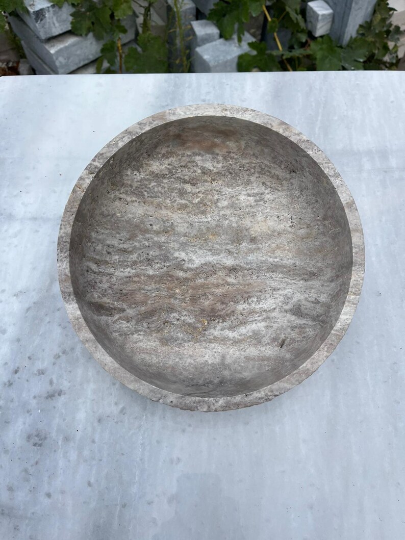 May include: A round, stone bowl with a textured, marbled appearance. The bowl is a light gray color with darker gray and brown veining throughout. The bowl's interior is smooth and the exterior has a rougher texture.