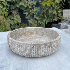 May include: A round, light-colored stone bowl with a textured, ribbed exterior. The bowl has a shallow interior and sits on a light-colored surface. The background features green foliage.