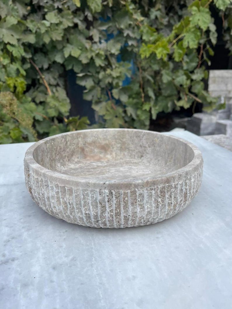 May include: A round, light-colored stone bowl with a textured, ribbed exterior. The bowl has a shallow interior and sits on a light-colored surface. The background features green foliage.