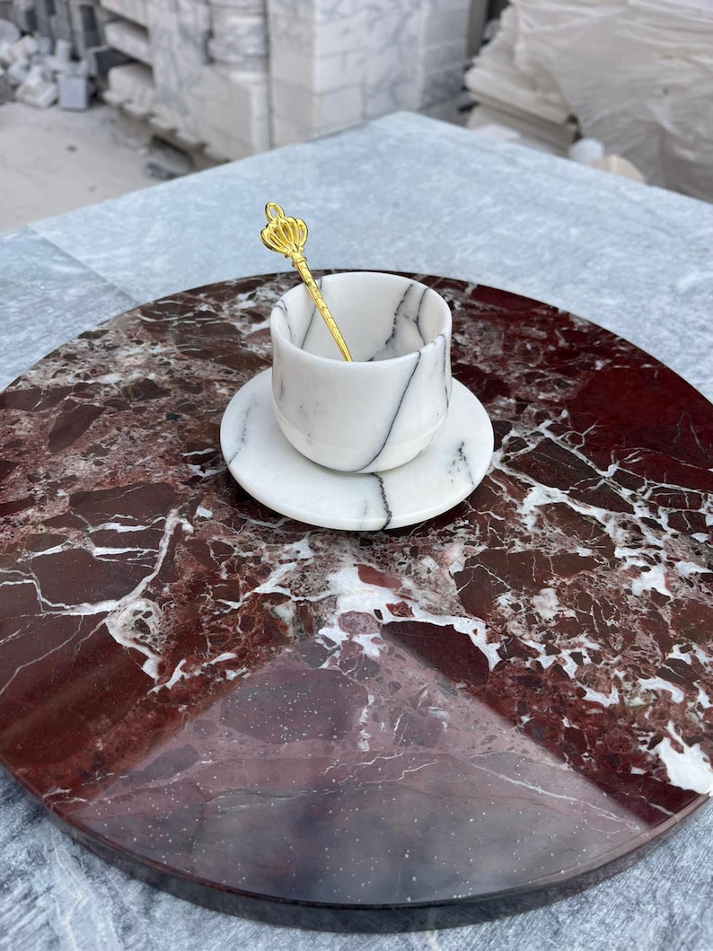 Op de afbeelding: Een witte marmeren kop en schotel set met zwarte adering, rustend op een ronde, rode marmeren dienblad. Een goudkleurige lepel met een kroonontwerp zit in de kop. De lade staat op een grijs oppervlak.