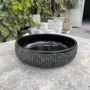 May include: A black, circular stone bowl with a textured, vertically lined exterior. The bowl has a smooth, glossy interior and rests on a grey stone surface. The bowl is likely for decorative or functional use.