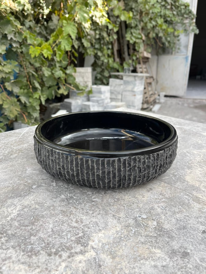 May include: A black, circular stone bowl with a textured, vertically lined exterior. The bowl has a smooth, glossy interior and rests on a grey stone surface. The bowl is likely for decorative or functional use.