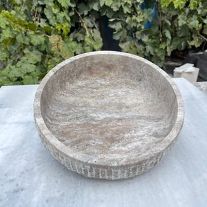 May include: A round, stone bowl with a textured surface and a light gray color. The bowl has a shallow depth and a wide rim with vertical grooves. The bowl is sitting on a white surface, with green foliage in the background.