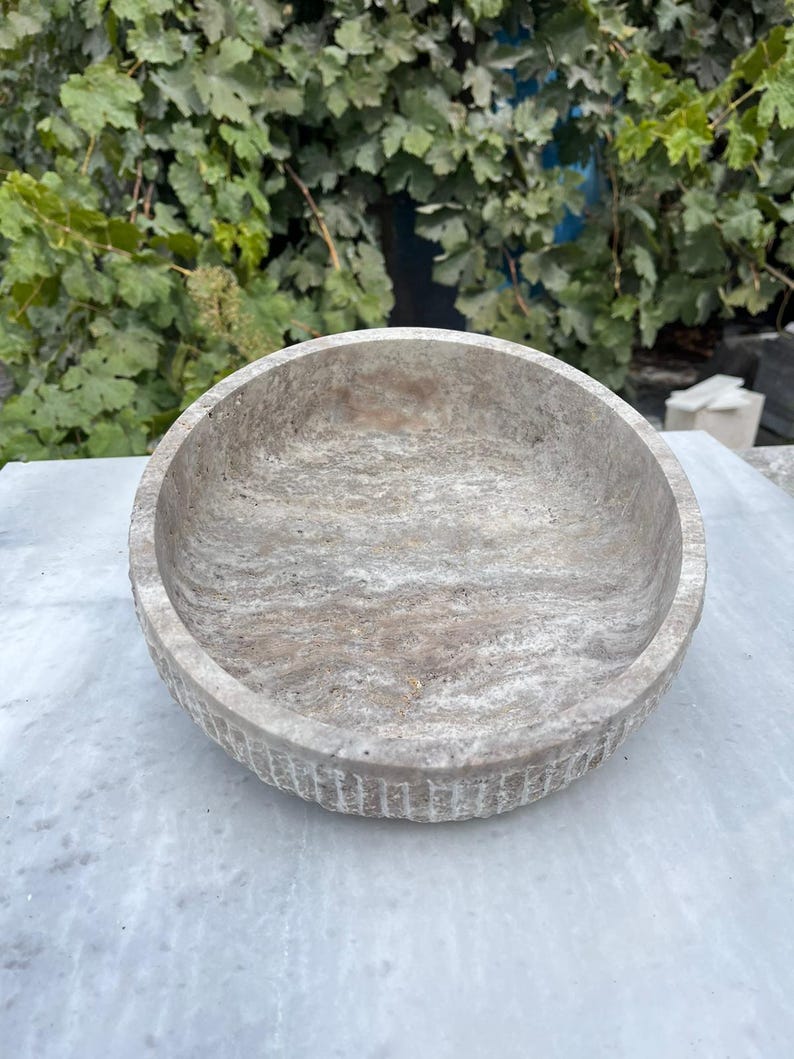 May include: A round, stone bowl with a textured surface and a light gray color. The bowl has a shallow depth and a wide rim with vertical grooves. The bowl is sitting on a white surface, with green foliage in the background.