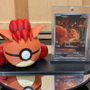 May include: An orange and red 3D-printed Vulpix figure next to a Vulpix Pokémon trading card in a protective case. The figure has a round body with a white and black circle detail and red accents. The card features a Vulpix illustration.
