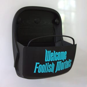May include: A matte black wall-mounted storage bin featuring a curved front panel with the text "Welcome Foolish Mortals" embossed in bright cyan lettering. The container has a rounded rectangular shape and a thin black bar across the top opening.