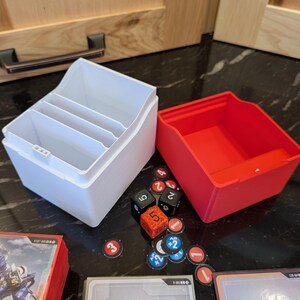 May include: Two plastic game organisers, one white with dividers and one red. Dice, tokens, and game cards are scattered around the organisers. The organisers are designed to hold board game components.