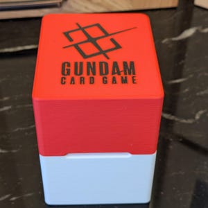 May include: A two-tone card game storage box. The top half is red, and the bottom half is white. The box has the words "GUNDAM CARD GAME" printed in black, along with a black graphic on the top.