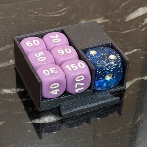 Puede incluir: Un soporte para dados negro impreso en 3D que contiene seis dados lavanda con n&uacute;meros blancos y un dado azul y blanco moteado. Los dados est&aacute;n dispuestos en dos compartimentos, sobre una superficie oscura y brillante.