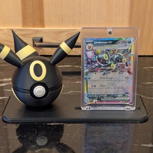 Puede incluir: Una figura de Umbreon negra y amarilla con una tarjeta a juego. La figura tiene detalles amarillos y un botón blanco. La tarjeta está en un soporte transparente y presenta una colorida ilustración de Umbreon.