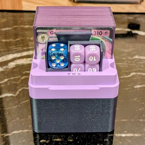 Puede incluir: Una caja de almacenamiento de cartas y dados impresa en 3D, de dos tonos. La parte superior y el interior son de color lavanda, mientras que la base es gris oscuro. En el interior hay cartas de juego, un dado azul y dados morados. La caja est&aacute; dise&ntilde;ada para guardar y organizar accesorios de juego.