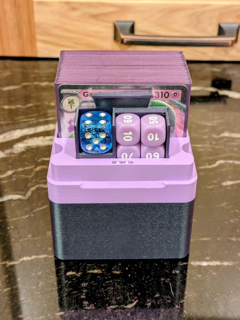 Puede incluir: Una caja de almacenamiento de cartas y dados impresa en 3D, de dos tonos. La parte superior y el interior son de color lavanda, mientras que la base es gris oscuro. En el interior hay cartas de juego, un dado azul y dados morados. La caja est&aacute; dise&ntilde;ada para guardar y organizar accesorios de juego.
