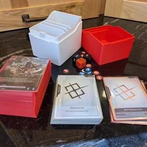 May include: A selection of trading card game accessories, including a white card storage box, a red storage box, dice, tokens, and stacks of cards with red edges. Several cards are visible, displaying game art and text.