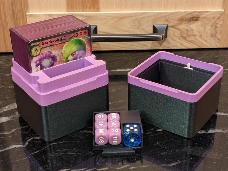 Puede incluir: Juego de almacenamiento de juegos de cartas de dos piezas. Los contenedores son negros con un borde morado. Un contenedor contiene una baraja de cartas con una carta Gardevoir EX visible. Una peque&ntilde;a bandeja contiene dados y contadores.