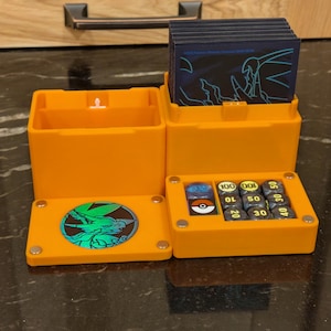 May include: An open, orange, three-compartment storage box for trading cards and dice. The box has a hinged lid with a circular emblem and a compartment for dice with numbers from 10 to 100. The box is on a dark, textured surface.