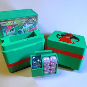 以下が含まれることがあります： 赤色のアクセントが付いた緑色のカード収納ボックス。トレーディングカードとサイコロが入っています。1つのボックスは開いており、「Iron Leaves GX」というテキストのカードが表示されています。もう一方のボックスには、蓋に赤と黒のグラフィックがあります。サイコロが見えます。