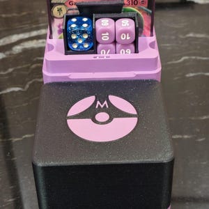 Puede incluir: Un soporte para cartas y dados bicolor impreso en 3D. La secci&oacute;n superior es lavanda y contiene una pila de cartas. La secci&oacute;n inferior es negra con un logotipo lavanda. Incluye un dado azul y dados morados.