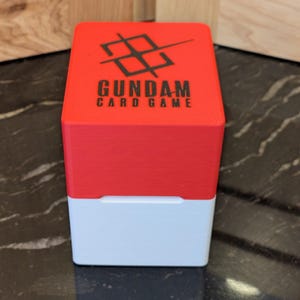 May include: A two-tone card game box with a red top and white base. The top features the text "GUNDAM CARD GAME" in black, with a black graphic above the text. The box is sitting on a black and grey surface.