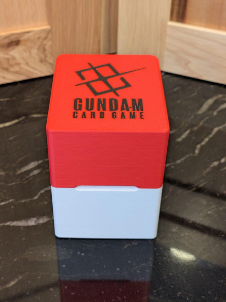 May include: A two-tone card game box with a red top and white base. The top features the text "GUNDAM CARD GAME" in black, with a black graphic above the text. The box is sitting on a black and grey surface.