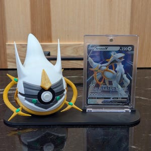 May include: A black display stand holding a white and gold Arceus figure and a Pokemon trading card in a protective case. The card features a white and blue Arceus V illustration. The figure has yellow and green accents.
