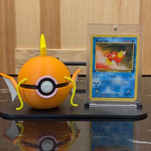 Puede incluir: Una figura de Magikarp naranja con aletas amarillas y un diseño de ojo blanco y negro, junto a una carta coleccionable de Magikarp en una funda protectora. La figura y la carta están sobre un soporte de exhibición negro.