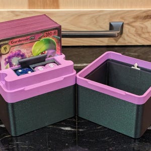 Puede incluir: Una caja de almacenamiento de cartas de dos tonos, con una base verde oscuro y una tapa rosa brillante. La tapa tiene un compartimento para dados y una pila de cartas. Se ve una sola carta, con un personaje Gardevoir.