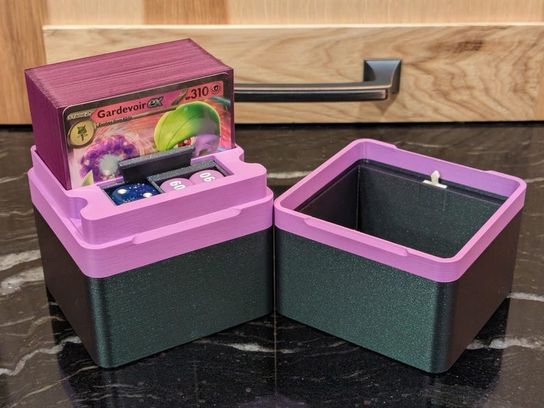 Puede incluir: Una caja de almacenamiento de cartas de dos tonos, con una base verde oscuro y una tapa rosa brillante. La tapa tiene un compartimento para dados y una pila de cartas. Se ve una sola carta, con un personaje Gardevoir.
