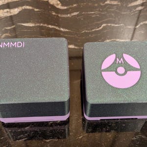 Puede incluir: Dos cajas cuadradas grises brillantes con bases negras y ribetes lavanda. Una caja tiene las letras "NMMDI" impresas en la parte superior. La otra caja tiene un logotipo lavanda con la letra "M". Las cajas est&aacute;n sobre una superficie reflectante.