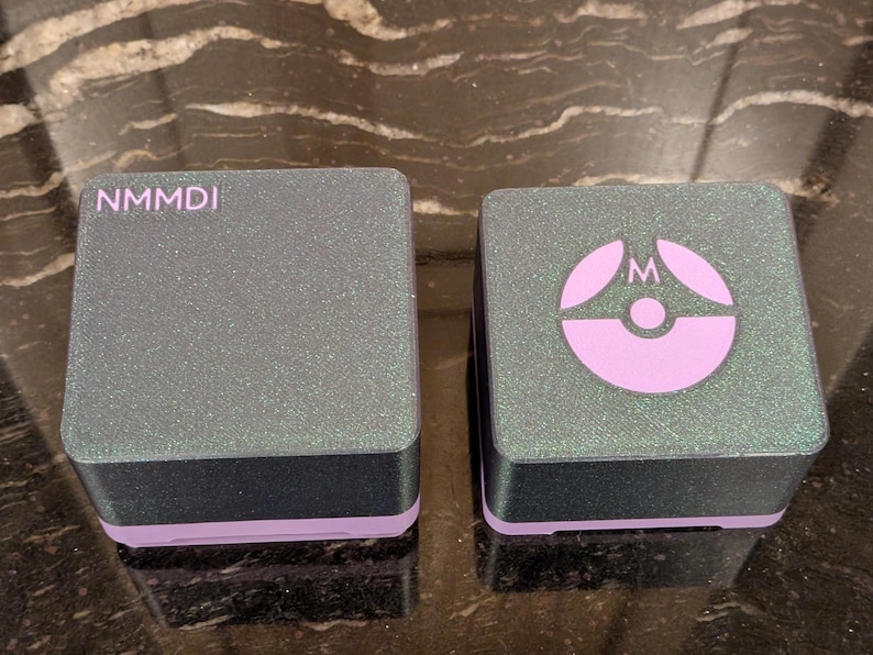 Puede incluir: Dos cajas cuadradas grises brillantes con bases negras y ribetes lavanda. Una caja tiene las letras "NMMDI" impresas en la parte superior. La otra caja tiene un logotipo lavanda con la letra "M". Las cajas est&aacute;n sobre una superficie reflectante.