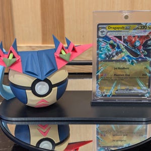 Könnte beinhalten: Ein 3D-gedrucktes Modell eines blau, rosa und beigen Charakters mit einer passenden Sammelkarte in einer Schutzhülle. Die Karte zeigt den Namen "Dragapult" und Spieldetails. Die Artikel stehen auf einer schwarzen, verspiegelten Oberfläche.