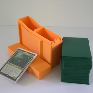 May include: An orange plastic card storage box with a removable lid, a single trading card encased in a clear protective sleeve, and a stack of solid dark green card sleeves positioned next to the open container on a plain white surface.