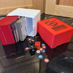 May include: A white card storage box with a red deck of cards and a red box with the text "GUNDAM CARD GAME". Dice and game tokens are scattered around the boxes. The game pieces are on a dark, reflective surface.