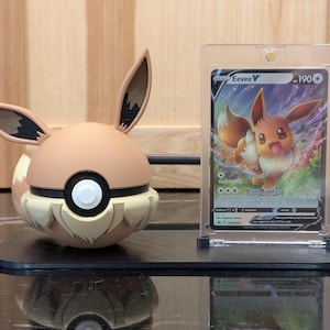 May include: A brown and tan Eevee figure with large ears and a white button detail, next to a protected Eevee V trading card. The items are displayed on a black stand, with a wooden background.
