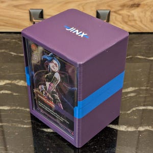 May include: A purple card storage box with the word "JINX" in white on the top. The box is secured with a blue band and contains a card featuring a character illustration. The card is labeled "Loose Cannon".
