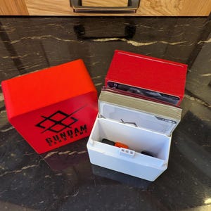 May include: A red card storage box with the text "GUNDAM CARD GAME" and a white card storage box with a red lid. The white box is open, revealing cards and small orange and black objects. A red box with cards is also visible.