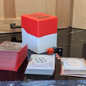 May include: A red and white card storage box with the word "KONNAN" on top, next to stacks of cards with red edges, dice, and game tokens. The cards have a geometric design.