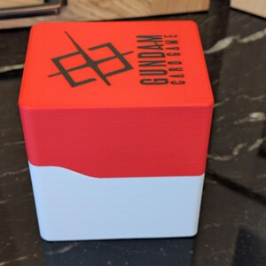 May include: A red and white card game box featuring the text "GUNDAM CARD GAME" and a black graphic. The box is rectangular with a curved design where the red and white sections meet.