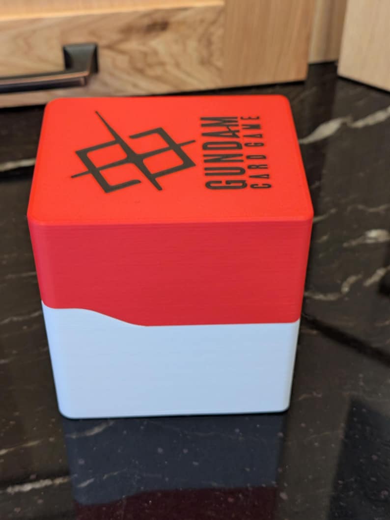 May include: A red and white card game box featuring the text "GUNDAM CARD GAME" and a black graphic. The box is rectangular with a curved design where the red and white sections meet.