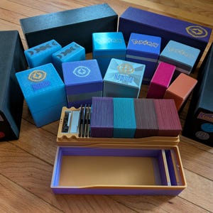 Peut inclure: Une collection de boîtes et de jeux de cartes colorés, notamment bleus, violets, roses et oranges. Certaines boîtes portent des logos et des textes, tels que "Lorcana" et "Neptune". Un plateau avec des accessoires de cartes est également visible.