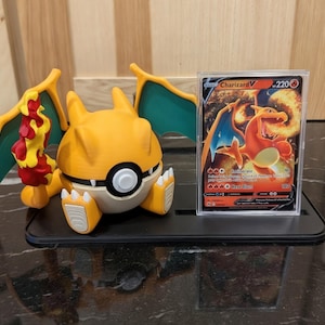 Puede incluir: Una figura de Charizard amarilla y blanca con alas verdes y llamas rojas y amarillas. Una carta coleccionable Charizard V se muestra en una funda transparente. Los artículos están sobre un soporte negro.