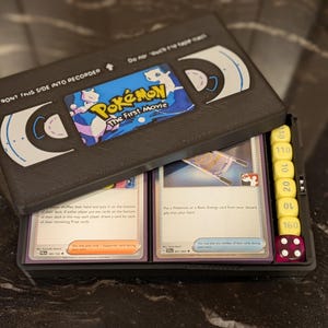 Peut inclure: Boîtier de rangement noir en forme de cassette VHS, avec le texte "FRONT THIS SIDE INTO RECORDER". Il contient des cartes à collectionner Pokémon, des dés et un autocollant de "Pokémon The First Movie".