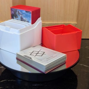 May include: A white card holder with compartments, holding red cards. A separate red box and a stack of cards with a geometric design are also present. The items are on a reflective surface.