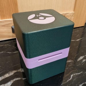 Puede incluir: Un contenedor verde oscuro en forma de cubo con una banda morada clara en el medio. La parte superior del contenedor tiene un logotipo blanco y morado. El contenedor est&aacute; sobre una superficie negra y gris.