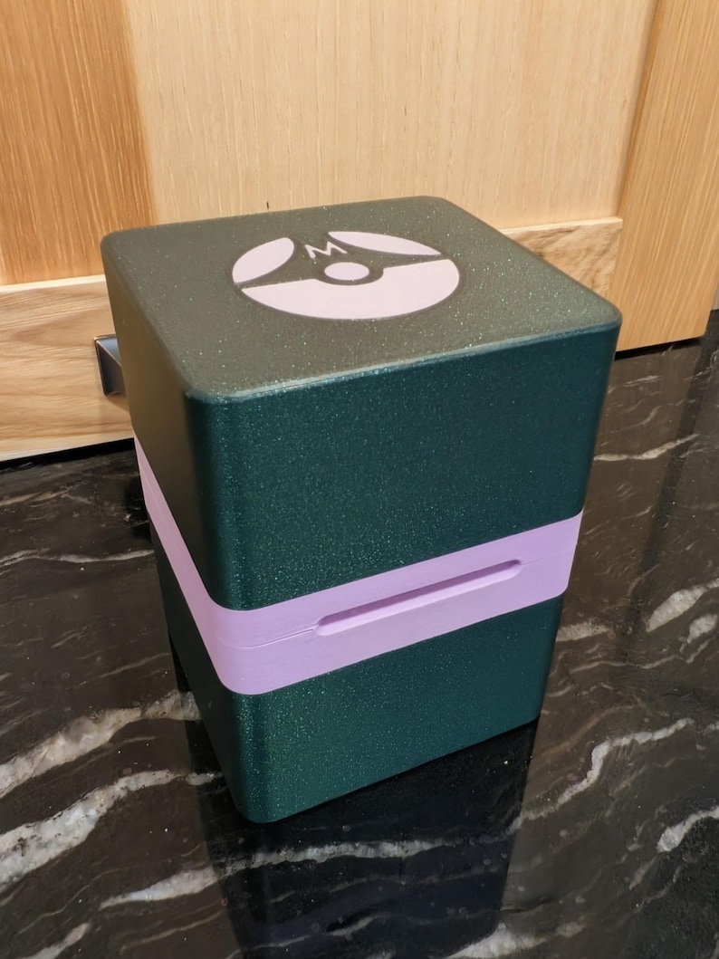 Puede incluir: Un contenedor verde oscuro en forma de cubo con una banda morada clara en el medio. La parte superior del contenedor tiene un logotipo blanco y morado. El contenedor est&aacute; sobre una superficie negra y gris.