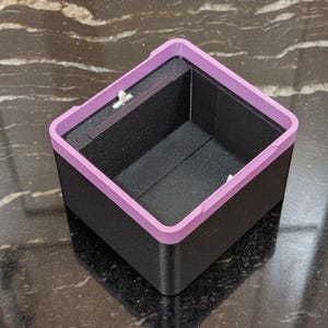 Puede incluir: Una caja cuadrada negra con un borde morado claro. El interior de la caja es negro. La caja est&aacute; sobre una superficie oscura y estampada.