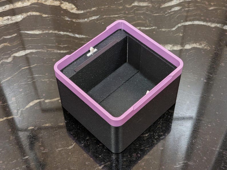 Puede incluir: Una caja cuadrada negra con un borde morado claro. El interior de la caja es negro. La caja est&aacute; sobre una superficie oscura y estampada.