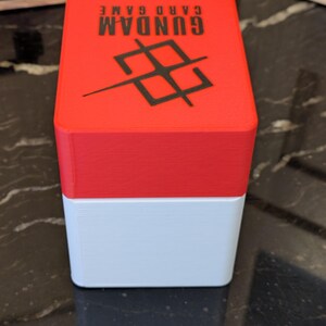 May include: A two-tone card game box, red on top and white on the bottom. The red lid features the text "GUNDAM CARD GAME" in black, along with a black geometric design. The box is designed to hold playing cards.