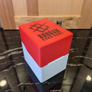 May include: A square card game box in red and white. The red lid is printed with the text "GUNDAM CARD GAME" in black, and a graphic. The box is made of plastic. The dimensions are not visible in the image.